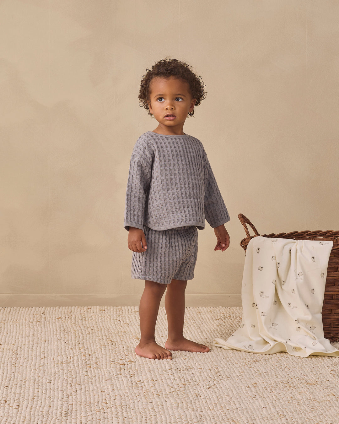 A toddler stands barefoot in a studio setting wearing a matching textured two-piece outfit in muted gray. The outfit consists of a long-sleeve top and shorts.