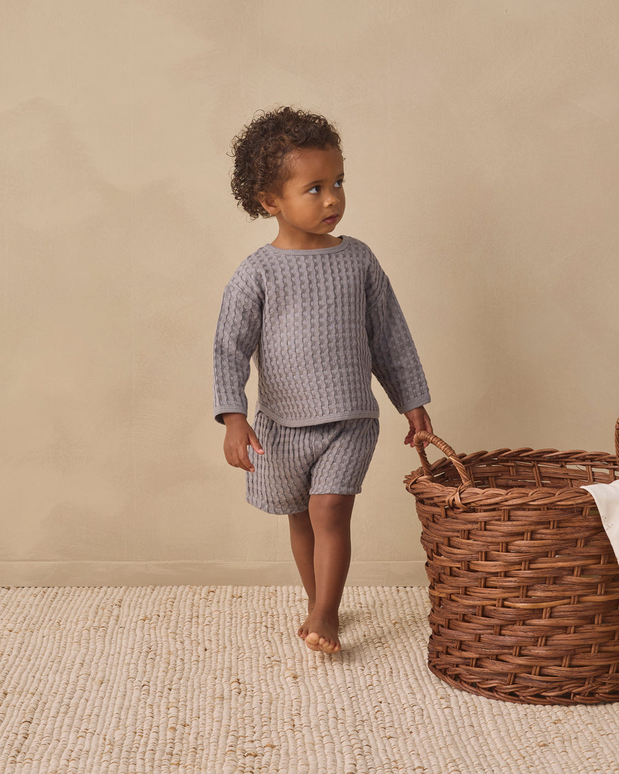 A toddler stands barefoot indoors wearing a matching textured knit two-piece outfit in a muted gray color. The outfit consists of a long-sleeve top and shorts.
