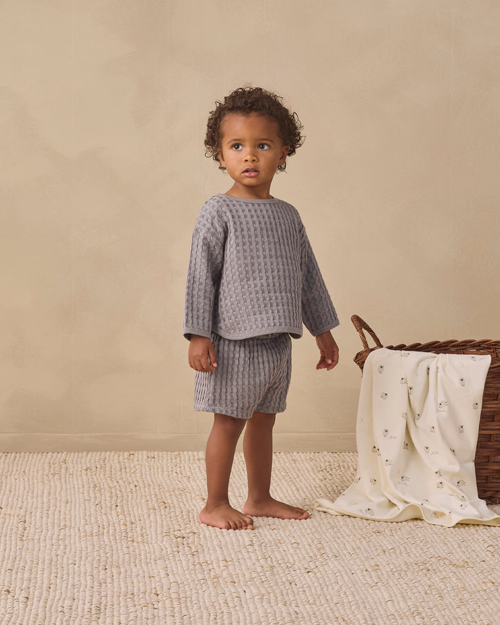 A toddler stands barefoot in a studio setting wearing a matching textured two-piece outfit in muted gray. The outfit consists of a long-sleeve top and shorts.