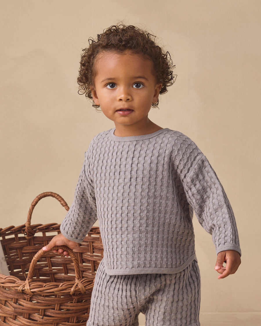 A toddler with curly brown hair wearing a light gray waffle knit long-sleeve top and matching pants, standing next to a wicker basket against a warm beige background.
