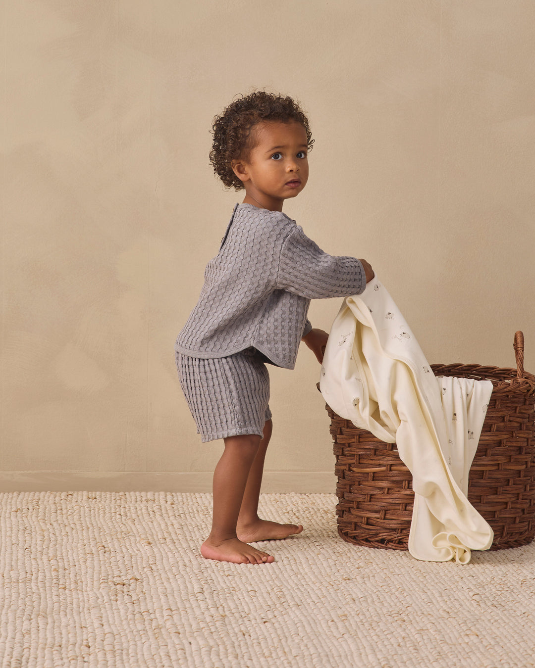 A toddler stands barefoot in a studio setting, wearing a textured light gray waffle set and holding a cream-colored blanket next to a wicker basket.