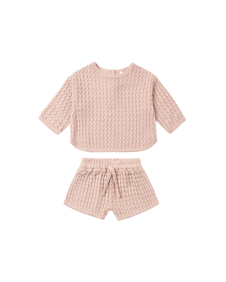 A two-piece baby outfit set in a dusty pink waffle-knit fabric, featuring a short-sleeve cropped top and matching shorts with an elastic waist and drawstring.