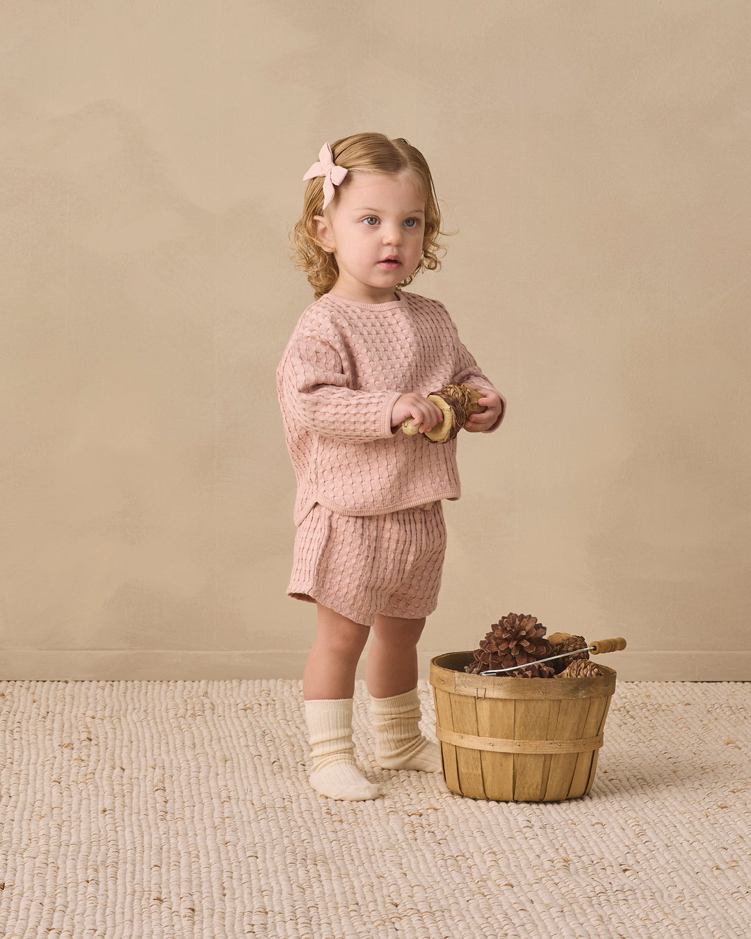 A toddler stands in a studio wearing a matching dusty pink waffle-knit long-sleeve top and shorts set, cream socks, and a pink bow in her hair. She is standing next to a small wooden basket on a textured cream rug against a warm beige background.