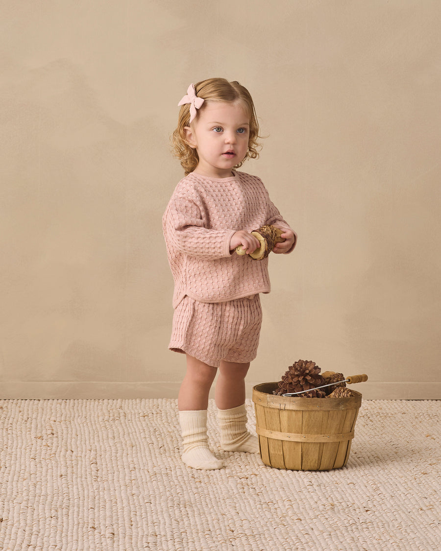 A toddler stands in a studio wearing a matching dusty pink waffle-knit long-sleeve top and shorts set, cream socks, and a pink bow in her hair. She is standing next to a small wooden basket on a textured cream rug against a warm beige background.