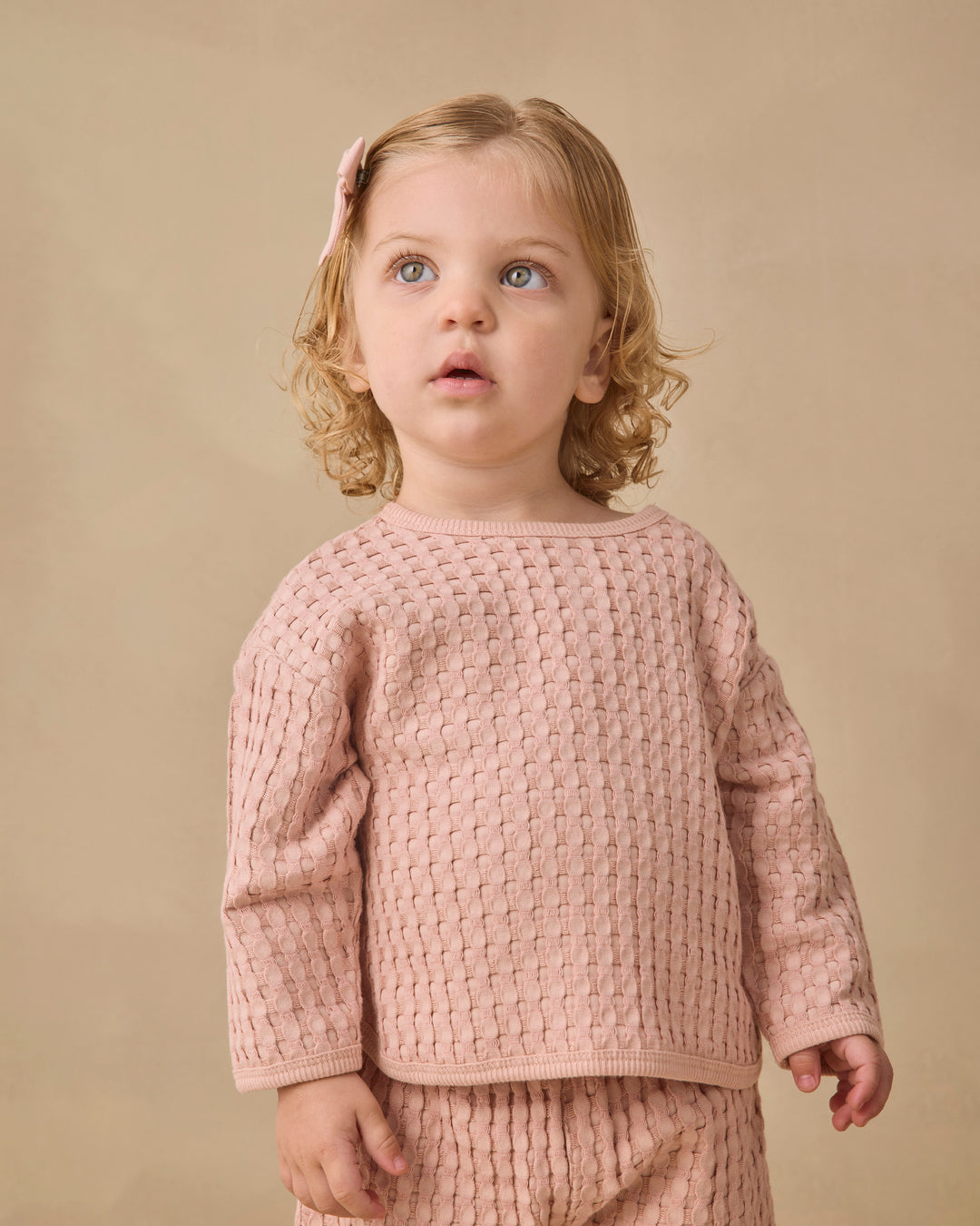 A young child with blonde curly hair wearing a dusty pink textured knit long-sleeve top and matching bottoms, standing against a solid beige background.
