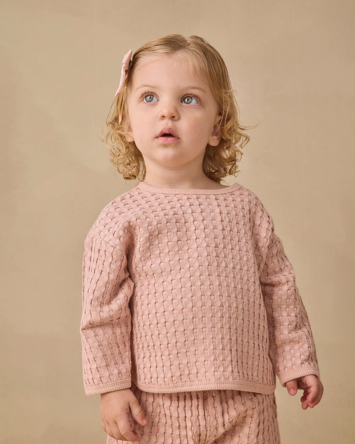 A young child with blonde curly hair wearing a dusty pink textured knit long-sleeve top and matching bottoms, standing against a solid beige background.
