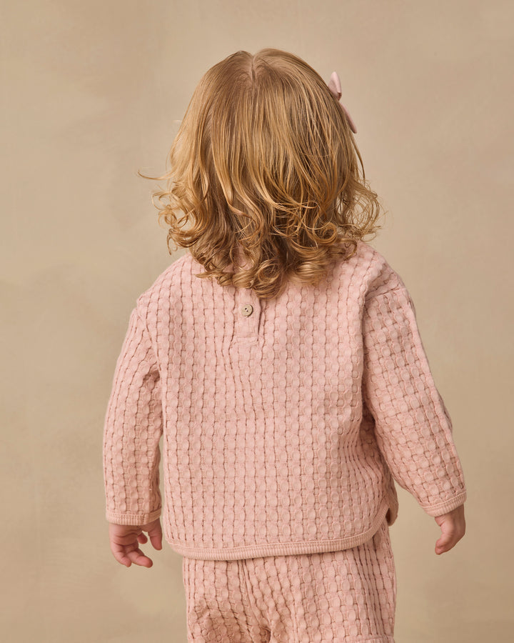 A toddler with blonde hair is shown from behind wearing a dusty pink textured knit two-piece outfit, consisting of a long-sleeve henley top with a button collar and matching knit pants, set against a solid beige background.