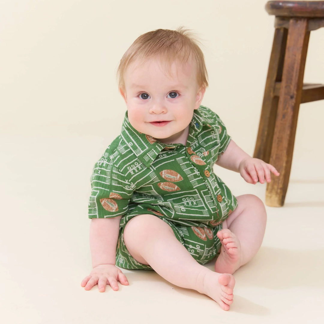 A baby sitting on a cream-colored floor wearing a green short-sleeve romper with a football field and football pattern, next to a wooden stool.