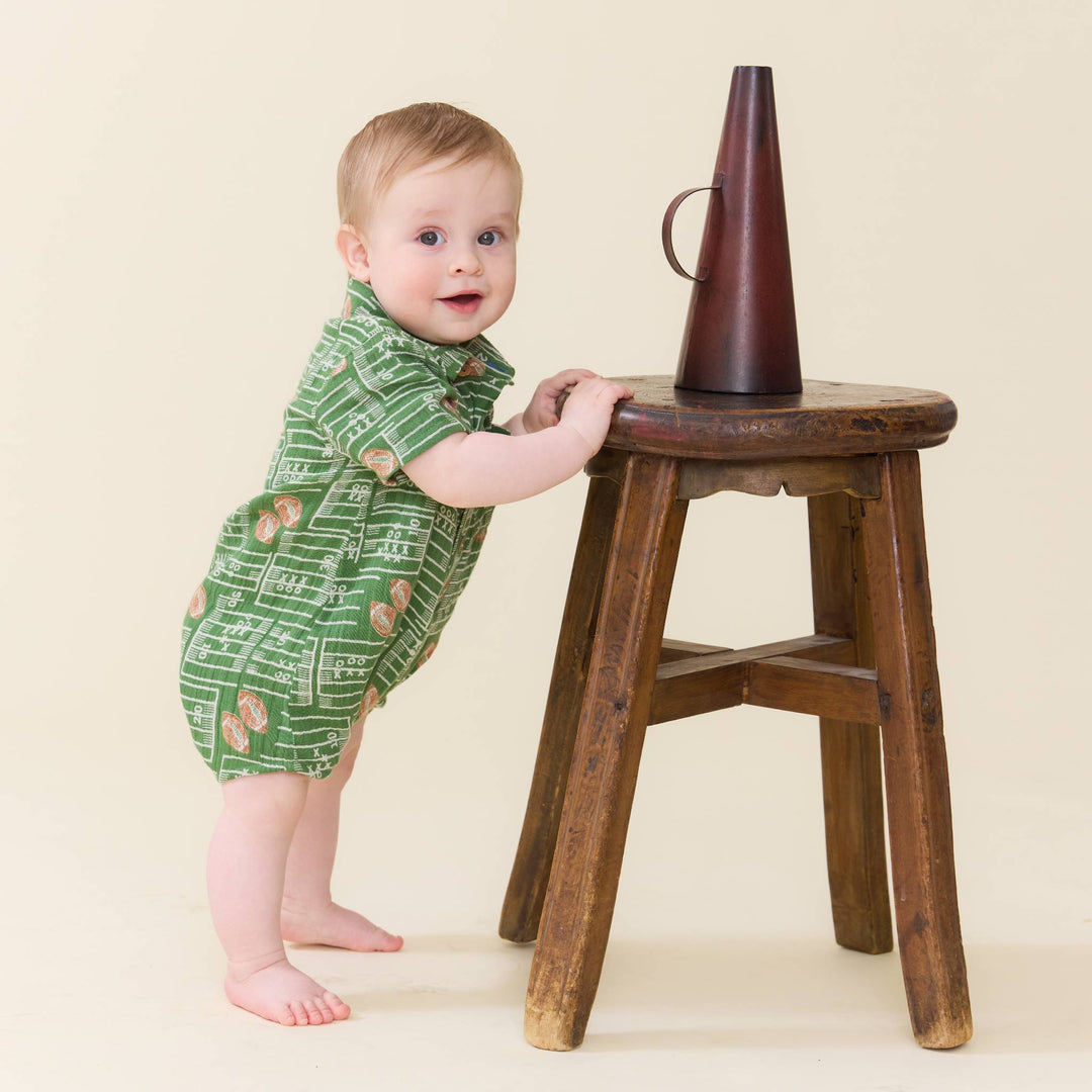 A baby standing on a wooden stool and holding onto it, wearing a green short-sleeve romper with a white musical notation print. A dark brown conical object sits on the stool.