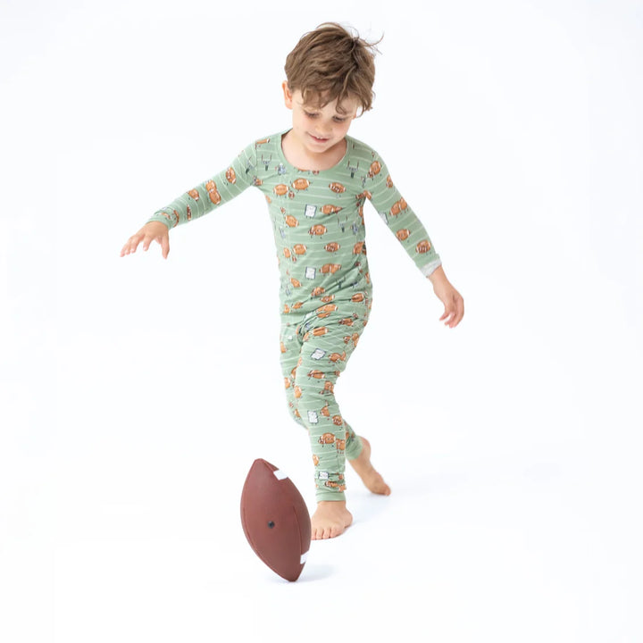 A young boy wearing a sage green long-sleeve pajama set with a cartoon pattern, playfully kicking a brown football on a seamless white background.