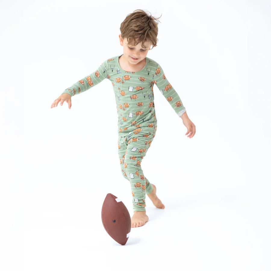 A young boy wearing a sage green long-sleeve pajama set with a cartoon pattern, playfully kicking a brown football on a seamless white background.