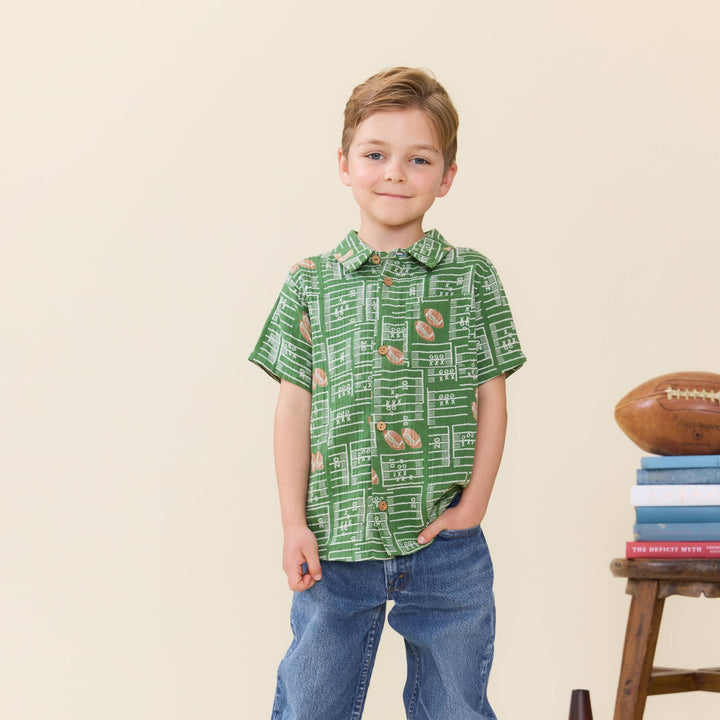 A young boy stands wearing a green short-sleeve button-up shirt with a chalkboard and football pattern, paired with blue jeans, against a warm beige background.