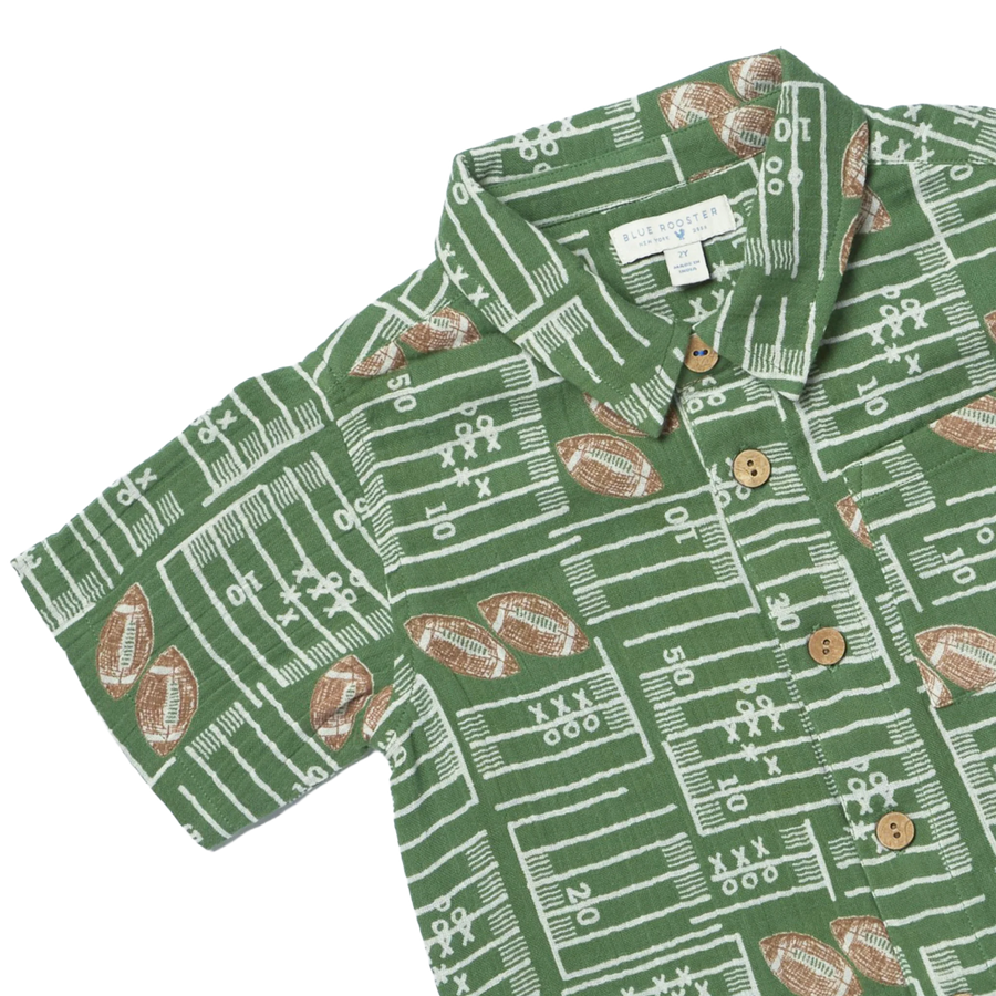 A green short-sleeve button-up shirt with a white football field and yard line pattern, accented with brown football motifs and brown buttons. The brand label reads 'The Happy Camp'.