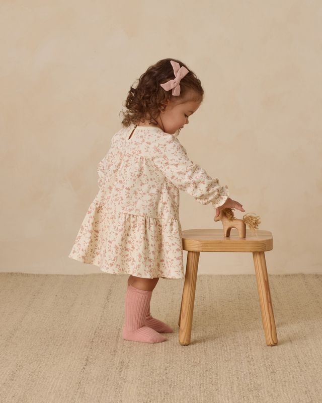 Child in a floral dress standing next to a wooden stool on a beige background