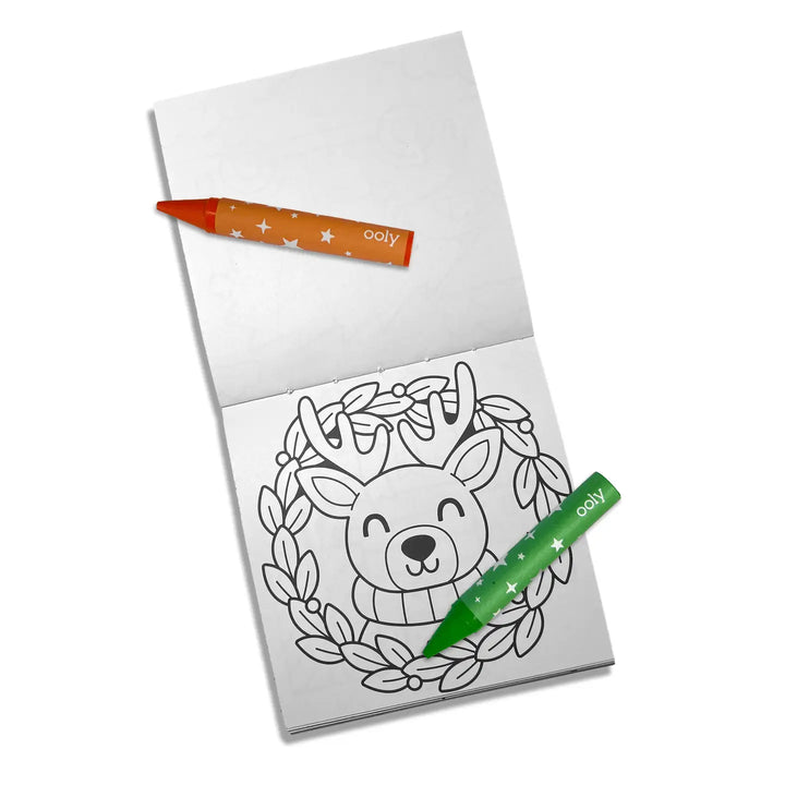 A small white coloring book or notepad on a white background, featuring a black-and-white reindeer face inside a wreath design on the lower page. Two crayons are placed on top: one orange with white stars and the word 'coly' near the tip, and one green with white stars and the same 'coly' branding.