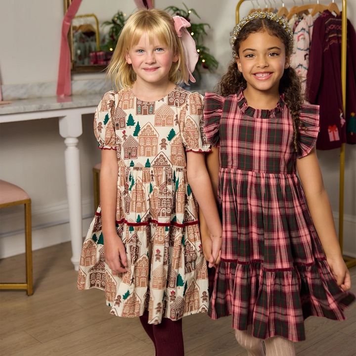 Two young girls standing indoors and holding hands, wearing long dresses with holiday-themed prints. The girl on the left wears a cream dress with gingerbread houses and trees, and the girl on the right wears a red and green plaid dress.
