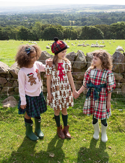 3 little girls holding hands in the Scotland dressed up in holiday outfits. one in plaid, one in a gingerbread dress and in a plaid skirt and holiday sweater