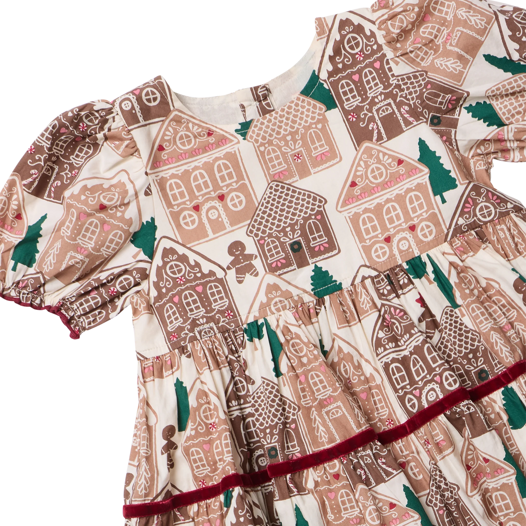 A close-up of a children's dress featuring a gingerbread house pattern in brown, beige, and pink, with green trees and a dark red ruffled trim along the skirt.