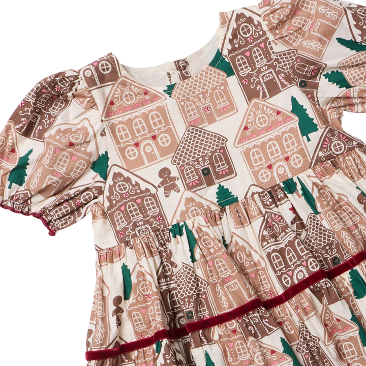 A close-up of a children's dress featuring a gingerbread house pattern in brown, beige, and pink, with green trees and a dark red ruffled trim along the skirt.