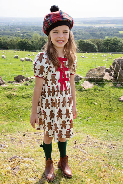 little girl modeling the gingerbread dress with 3 red bows at the top of the dress. the dress is short sleeves and hits at the knee. 