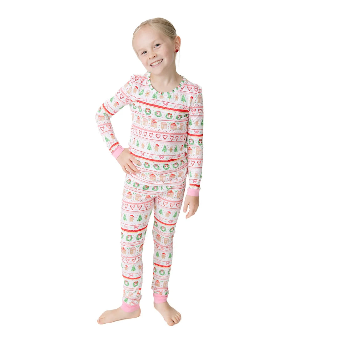 A young girl stands barefoot against a plain white background wearing a long-sleeve pajama set with a festive holiday pattern in red, green, and blue on a white base.