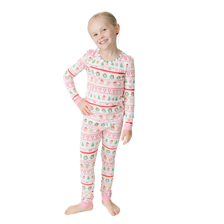 A young girl stands barefoot against a plain white background wearing a long-sleeve pajama set with a festive holiday pattern in red, green, and blue on a white base.