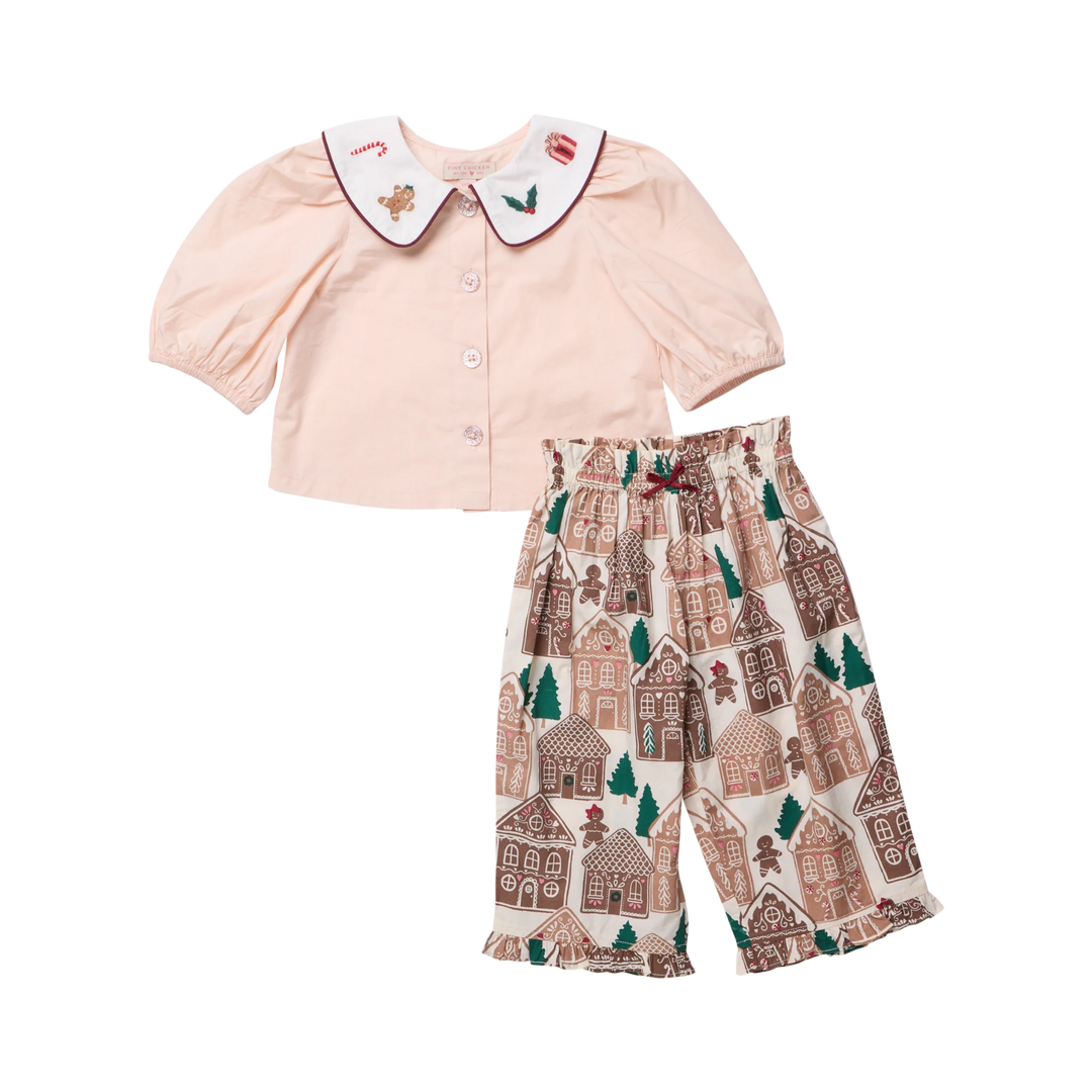 A two-piece baby outfit set featuring a pale pink short-sleeve top with a white Peter Pan collar decorated with small embroidered motifs, paired with cream-colored pants printed with a brown and green gingerbread house pattern.