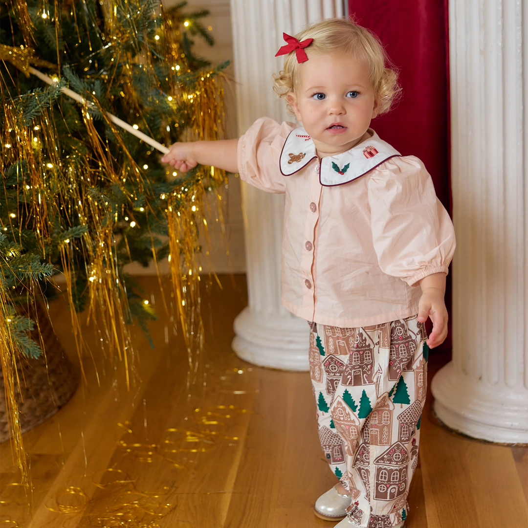 A toddler girl wearing a pale pink holiday-themed button-up shirt with a white embroidered collar and matching gingerbread-patterned pants, standing in a decorated room next to a Christmas tree with gold tinsel.