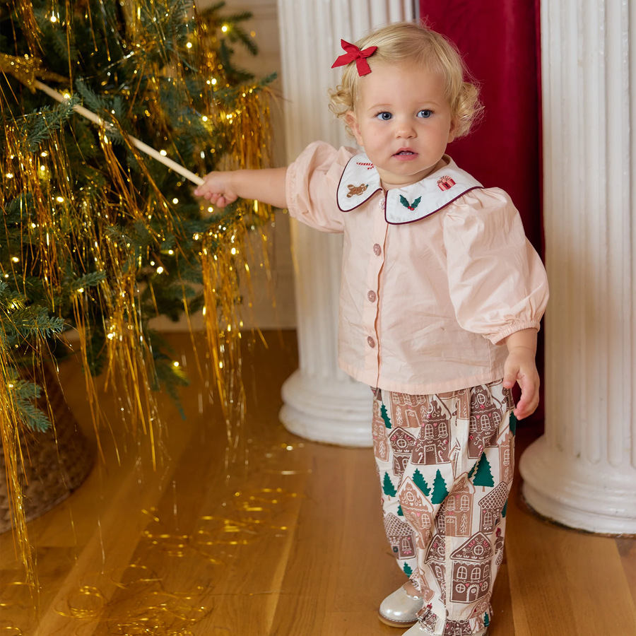 A toddler girl wearing a pale pink holiday-themed button-up shirt with a white embroidered collar and matching gingerbread-patterned pants, standing in a decorated room next to a Christmas tree with gold tinsel.