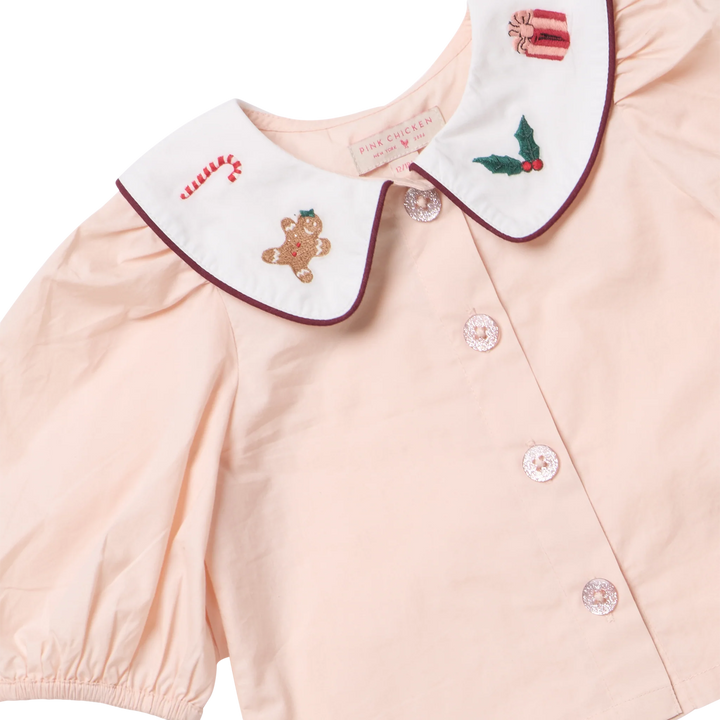 A close-up of a pale peach-pink short-sleeve blouse with a large white Peter Pan collar decorated with Christmas-themed embroidery, including a gingerbread man, candy cane, gift box, and holly. The blouse has a button front and is shown on a clean white background.
