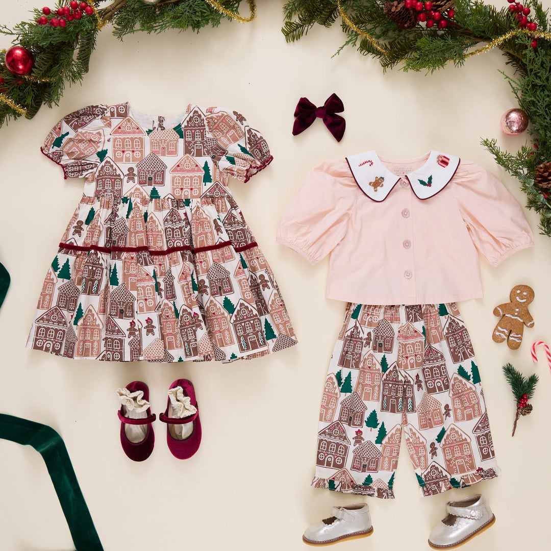 A flat-lay of a baby girl's holiday outfit set on a cream background decorated with Christmas garlands. The set includes a pink long-sleeve top with a white Peter Pan collar, matching cream pants with a gingerbread house pattern, a white tiered dress with a gingerbread house pattern, burgundy Mary Jane shoes, and a burgundy bow.
