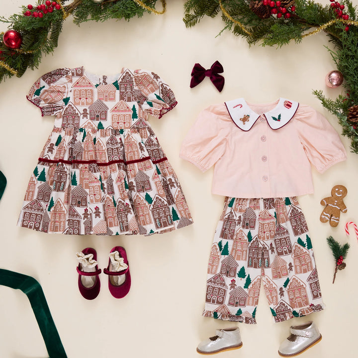 A flat-lay of a baby girl's holiday outfit set on a cream background decorated with Christmas garlands. The set includes a pink long-sleeve top with a white Peter Pan collar, matching cream pants with a gingerbread house pattern, a white tiered dress with a gingerbread house pattern, burgundy Mary Jane shoes, and a burgundy bow.