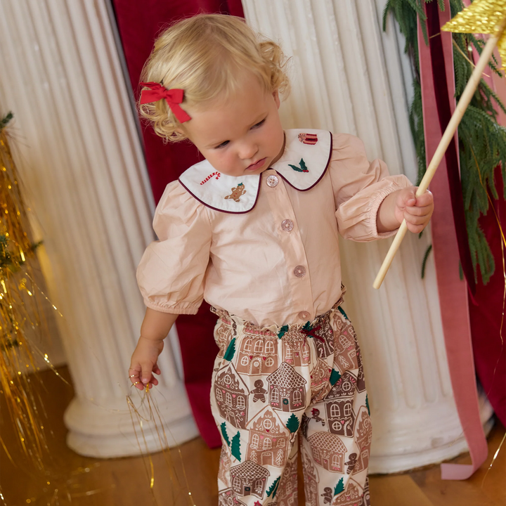 A toddler girl wearing a pale peach puff-sleeve top with a white embroidered collar and matching holiday-print pants, standing in a decorated indoor setting with columns and gold tinsel.