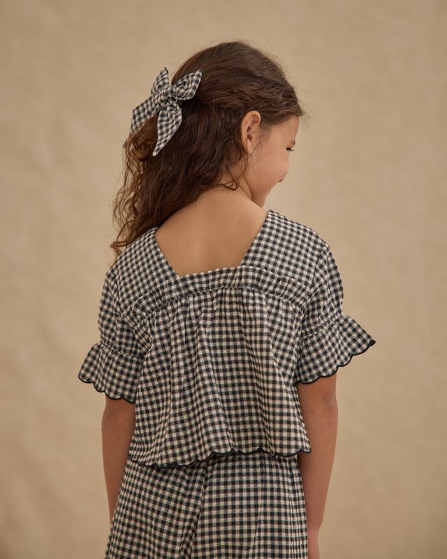 Young girl wearing a black and white gingham from the back- the top has an outfit with ruffled sleeves against a beige background