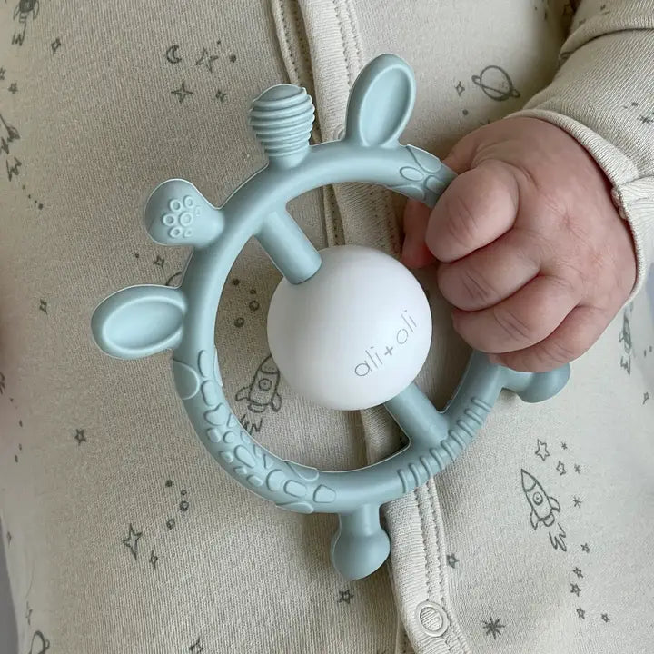 Baby hand holding a blue teething ring with a white ball, branded 'ali+oli', against a neutral background.