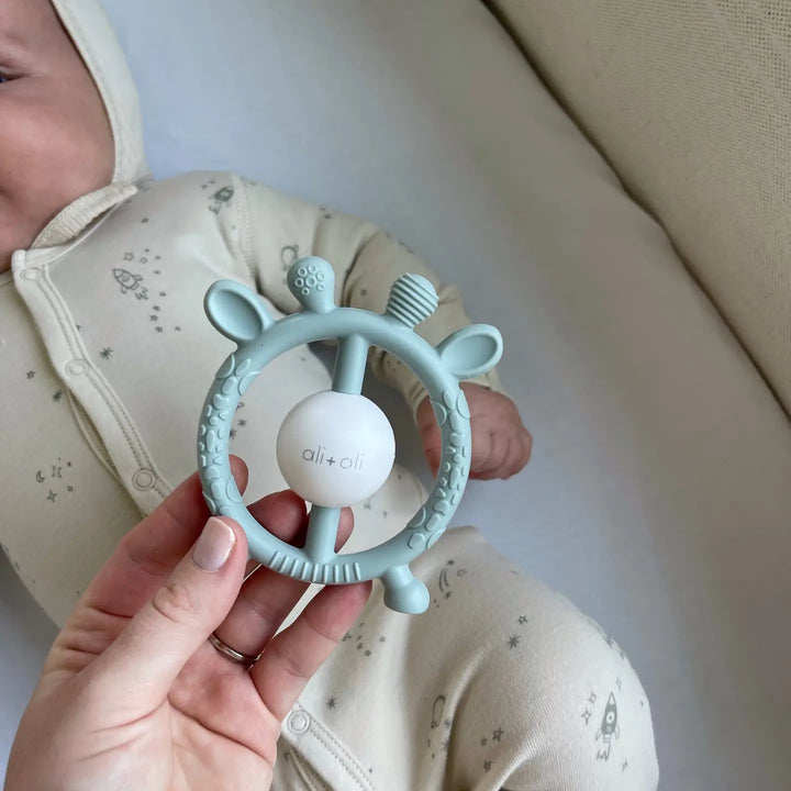 Baby holding a light blue teething ring with a white ball, against a neutral background.