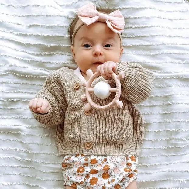 Baby wearing a knitted cardigan and floral pants, holding a teething ring on a textured white background