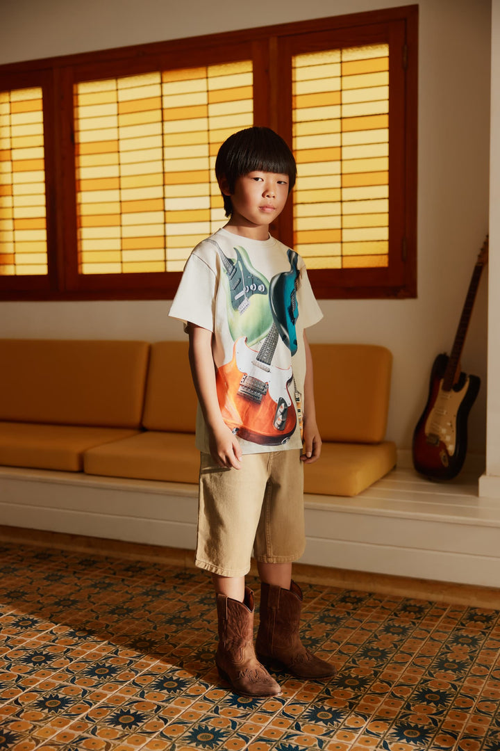 Child wearing a graphic t-shirt and beige shorts standing in a room with a guitar and window with wooden frames.