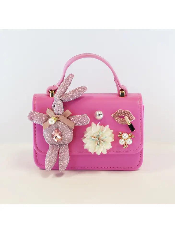 pink handbag with flap and snap closure. glitter pink bunny and flower and charms attached to the front of the bag. handle and gold chain that is detachable are options for carrying the bag
