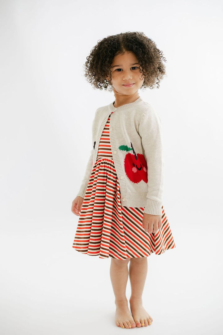 A young child with curly dark hair stands barefoot against a plain white studio background, wearing a cream knit cardigan over a red and white striped dress with a red apple applique on the front.