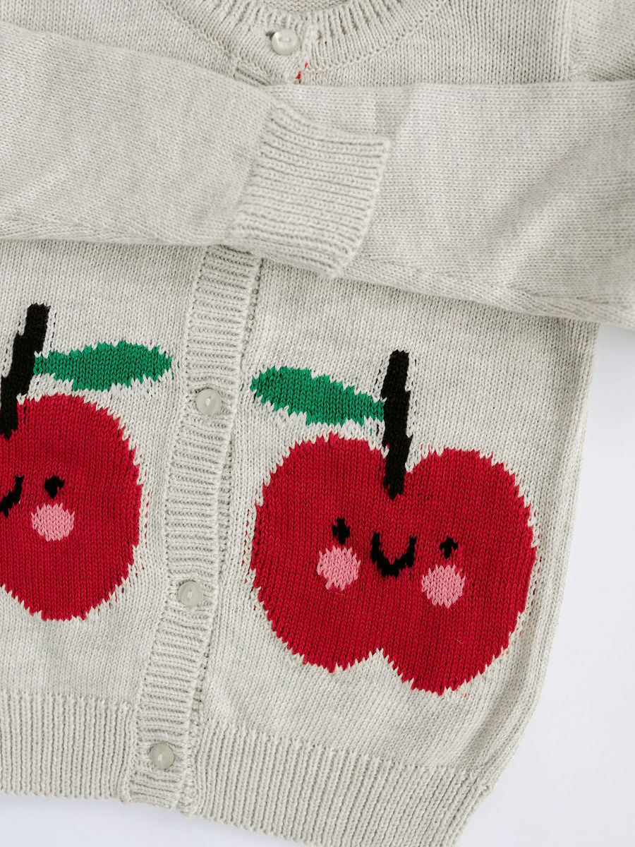 Close-up of a light beige knit cardigan sweater featuring two large red apple motifs with green leaves and smiling faces knitted onto the front.