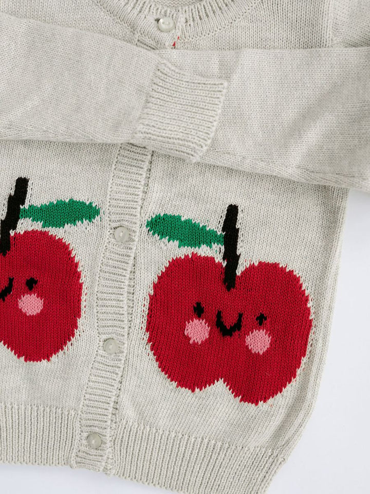 Close-up of a light beige knit cardigan sweater featuring two large red apple motifs with green leaves and smiling faces knitted onto the front.