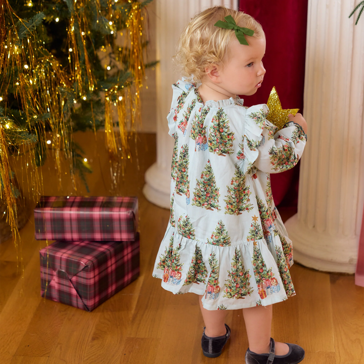 A toddler girl wearing a light blue ruffled dress with a Christmas tree print stands in a holiday-themed room next to a decorated Christmas tree and wrapped gifts.