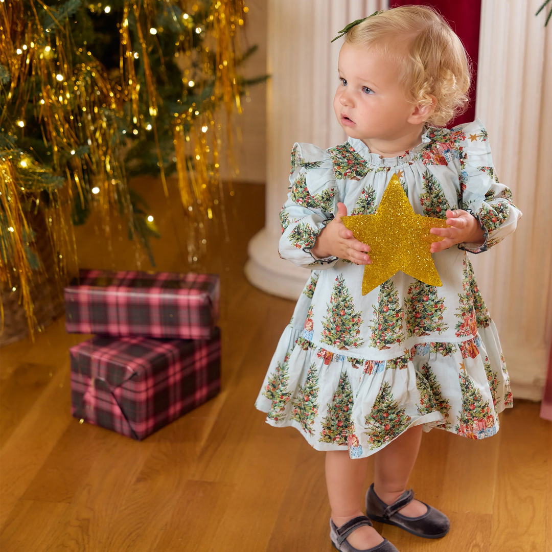 A toddler girl wearing a white long-sleeve dress with a Christmas tree print stands in a holiday room holding a glittery gold star ornament. A Christmas tree with gold tinsel, wrapped gifts, and a wooden floor are visible in the background.