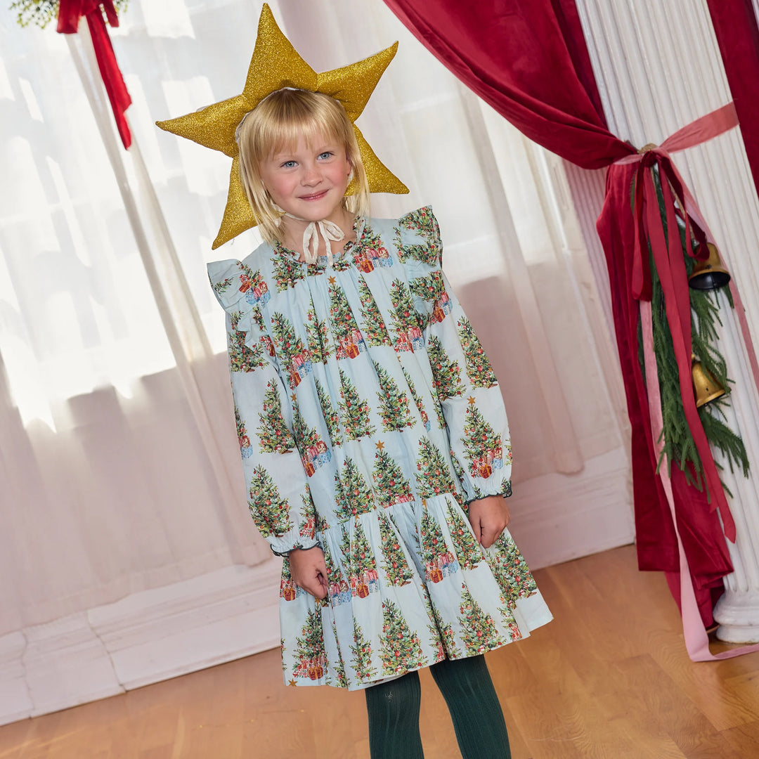 A young girl stands indoors wearing a light blue long-sleeve dress with an allover Christmas tree print and a large gold star headpiece. The room has white curtains, a red velvet curtain, and a wooden floor.