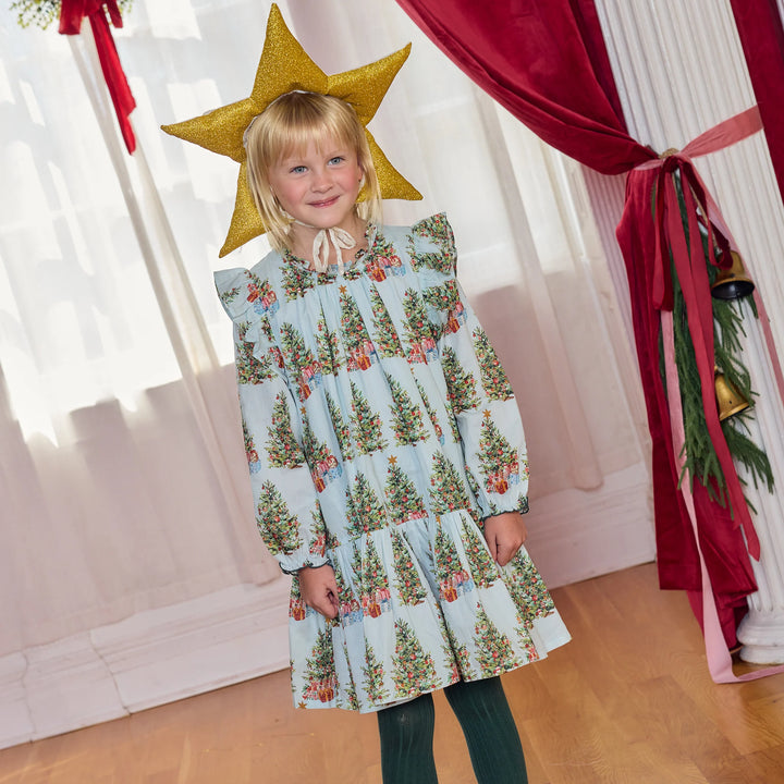 A young girl stands indoors wearing a light blue long-sleeve dress with an allover Christmas tree print and a large gold star headpiece. The room has white curtains, a red velvet curtain, and a wooden floor.