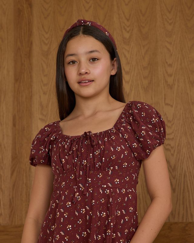 Young girl wearing a burgundy dress with a subtle pattern against a wooden paneled background