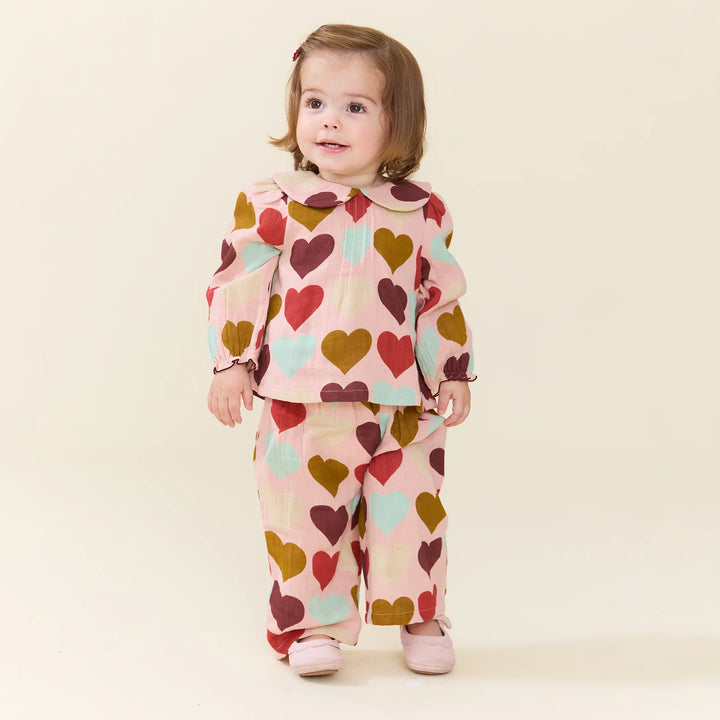 A toddler girl wearing a matching two-piece pajama set with a heart pattern, standing against a solid cream background.