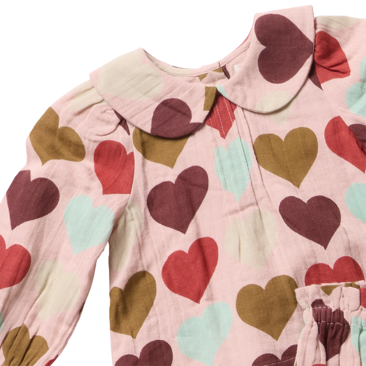 A close-up of a children's long-sleeve top with a Peter Pan collar, featuring a pink base fabric covered in a pattern of large hearts in red, brown, and light blue.