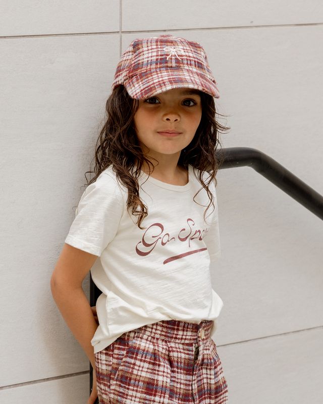 Child wearing a white t-shirt with text and plaid hat and pants against a light gray wall.